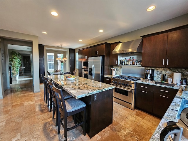 kitchen with light stone counters, a center island, stainless steel appliances, dark brown cabinets, and wall chimney exhaust hood