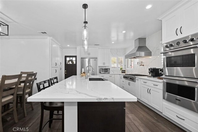 kitchen with appliances with stainless steel finishes, white cabinetry, a breakfast bar area, a kitchen island with sink, and wall chimney exhaust hood