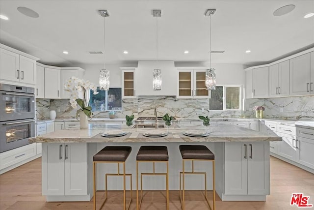 kitchen featuring light stone countertops, double oven, a center island with sink, and decorative light fixtures