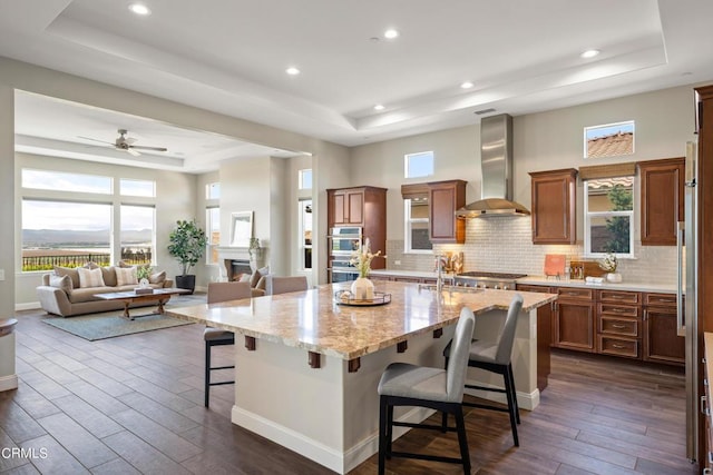 kitchen with wall chimney range hood, a tray ceiling, a kitchen island with sink, a kitchen breakfast bar, and dark hardwood / wood-style flooring