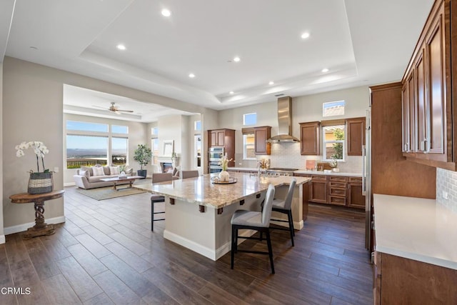 kitchen with a spacious island, a kitchen breakfast bar, a tray ceiling, and wall chimney exhaust hood