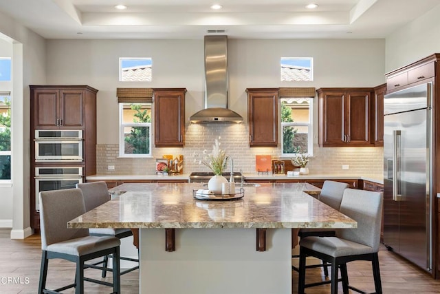 kitchen with a kitchen bar, wall chimney range hood, a center island with sink, and stainless steel appliances