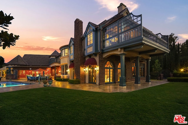 back house at dusk featuring a patio area, a lawn, and a balcony