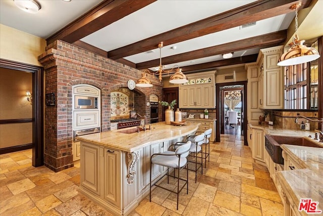 kitchen with cream cabinetry, an island with sink, pendant lighting, light stone counters, and beamed ceiling