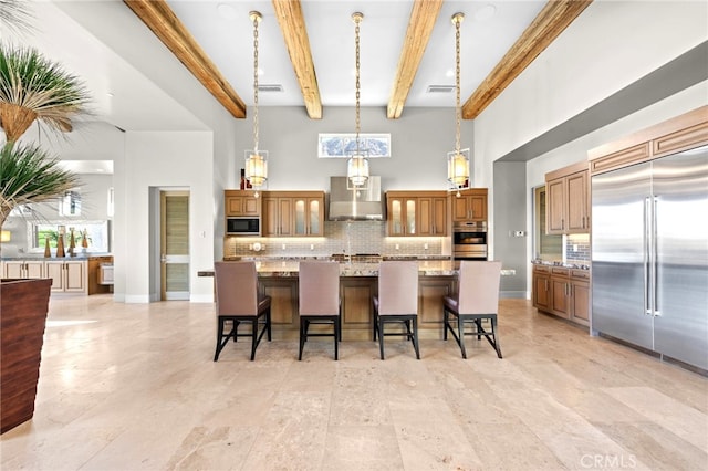 kitchen with wall chimney range hood, a spacious island, built in appliances, light stone countertops, and a kitchen bar