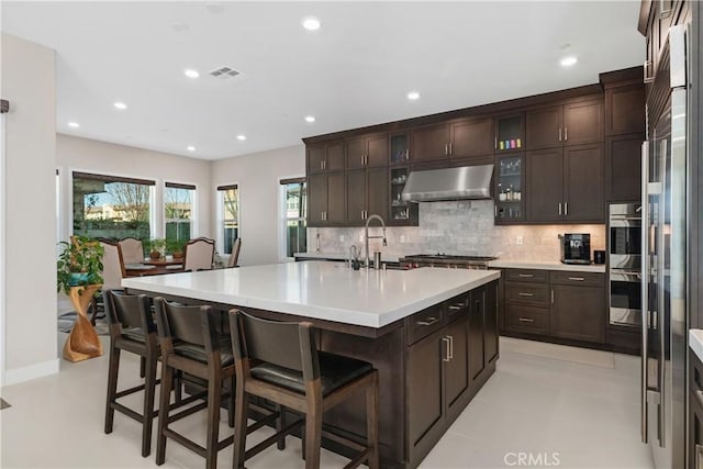 kitchen with sink, a breakfast bar, dark brown cabinetry, ventilation hood, and a center island with sink