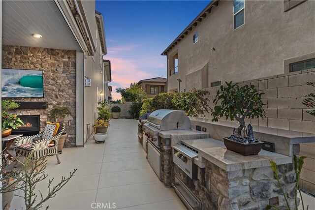 patio terrace at dusk featuring exterior kitchen, area for grilling, and an outdoor stone fireplace