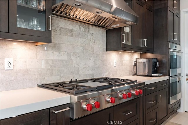 kitchen featuring stainless steel appliances, dark brown cabinetry, exhaust hood, light countertops, and decorative backsplash