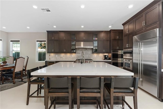 kitchen featuring stainless steel appliances, a breakfast bar, wall chimney range hood, and a large island with sink