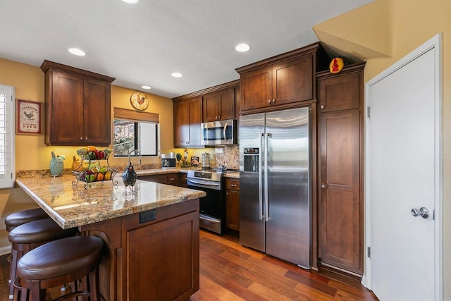 kitchen featuring appliances with stainless steel finishes, kitchen peninsula, a breakfast bar, light stone counters, and dark hardwood / wood-style floors