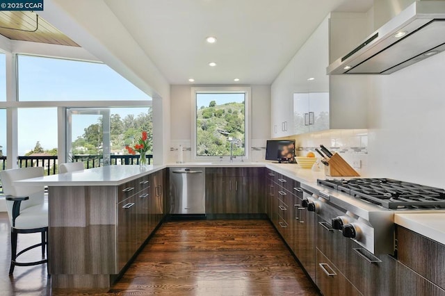 kitchen with wall chimney range hood, stainless steel appliances, white cabinets, a kitchen bar, and kitchen peninsula