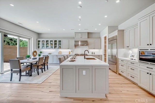 kitchen with a center island with sink, light countertops, glass insert cabinets, stainless steel built in fridge, and wall chimney range hood