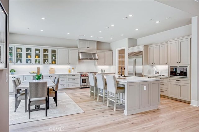 kitchen featuring a center island with sink, light countertops, glass insert cabinets, ventilation hood, and built in appliances