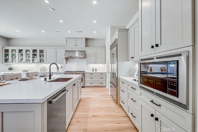 kitchen with built in appliances, a sink, light countertops, wall chimney exhaust hood, and glass insert cabinets