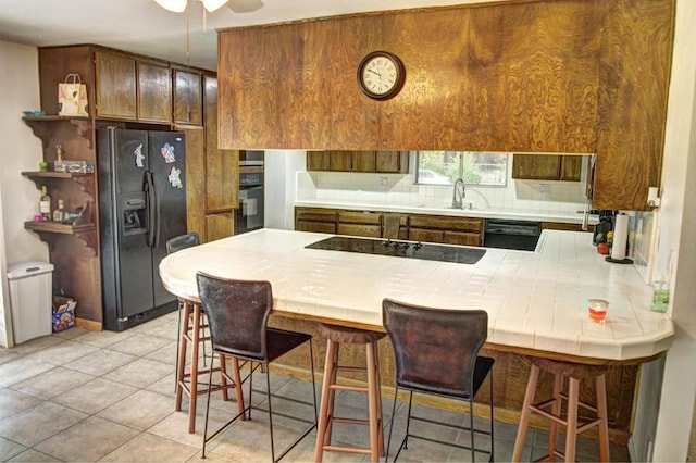 kitchen featuring a breakfast bar, tile countertops, black appliances, light tile patterned floors, and kitchen peninsula