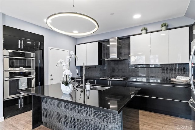 kitchen featuring appliances with stainless steel finishes, wall chimney exhaust hood, sink, light wood-type flooring, and white cabinetry