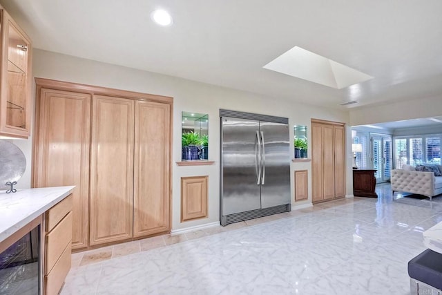 kitchen with built in refrigerator, a skylight, light stone countertops, light brown cabinetry, and beverage cooler