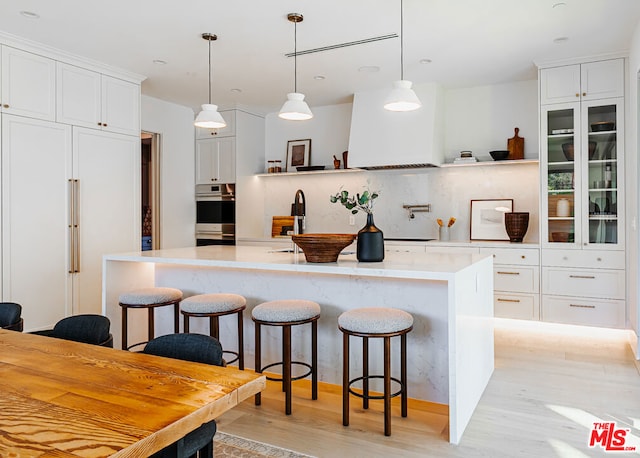 kitchen with a kitchen bar, light hardwood / wood-style floors, hanging light fixtures, white cabinets, and an island with sink