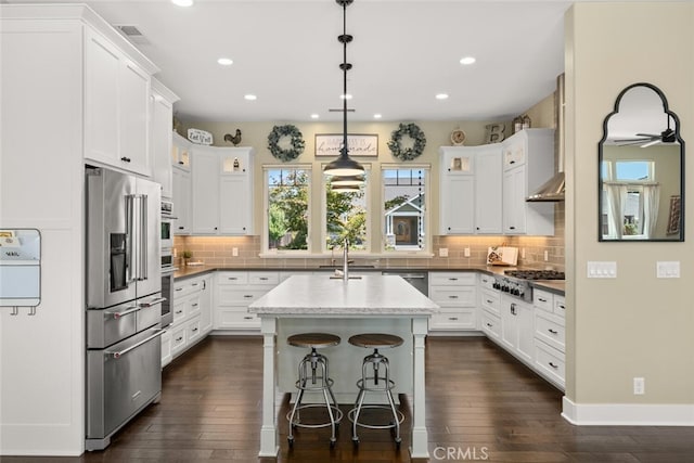 kitchen featuring white cabinets, appliances with stainless steel finishes, decorative light fixtures, dark stone counters, and a center island with sink