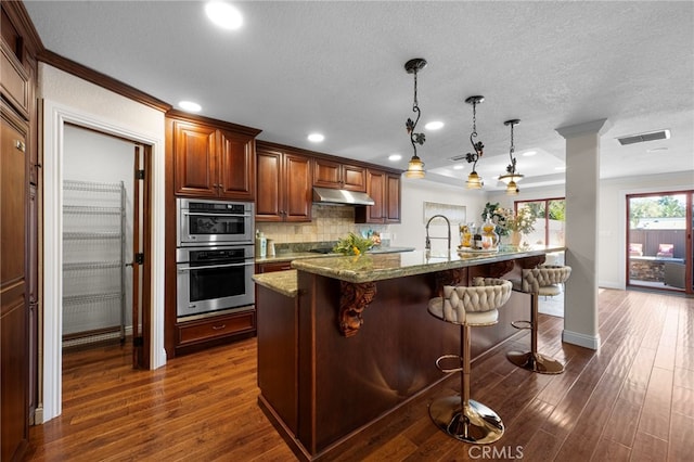 kitchen featuring decorative light fixtures, backsplash, light stone countertops, a kitchen island with sink, and a kitchen bar