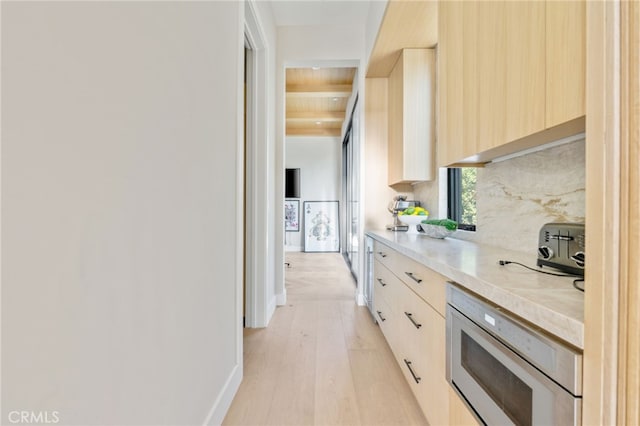 kitchen featuring stainless steel microwave, light brown cabinetry, tasteful backsplash, beam ceiling, and light hardwood / wood-style flooring