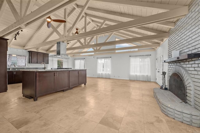 kitchen with island exhaust hood, a fireplace, a kitchen island with sink, beamed ceiling, and dark brown cabinetry