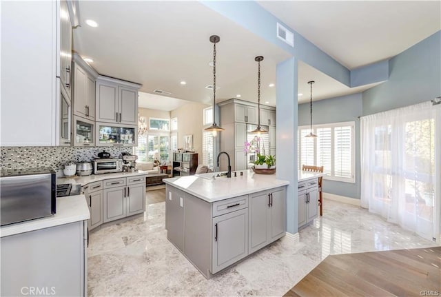 kitchen with gray cabinets, a center island with sink, backsplash, and decorative light fixtures