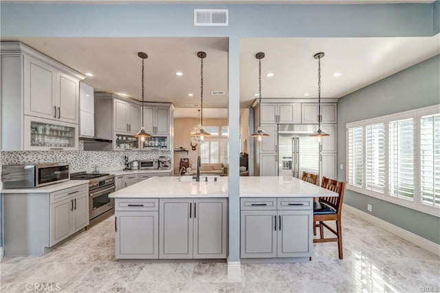 kitchen with gray cabinets, appliances with stainless steel finishes, hanging light fixtures, tasteful backsplash, and a center island with sink