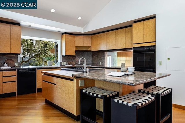 kitchen with vaulted ceiling, light wood-type flooring, dark stone countertops, a center island with sink, and black appliances