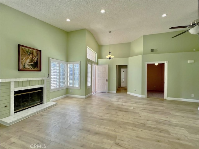 unfurnished living room featuring a textured ceiling, light wood-type flooring, a towering ceiling, a fireplace, and ceiling fan with notable chandelier