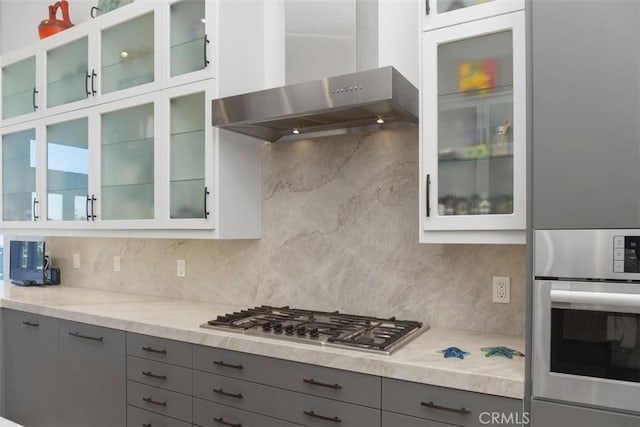 kitchen with wall chimney range hood, stainless steel appliances, gray cabinetry, and backsplash