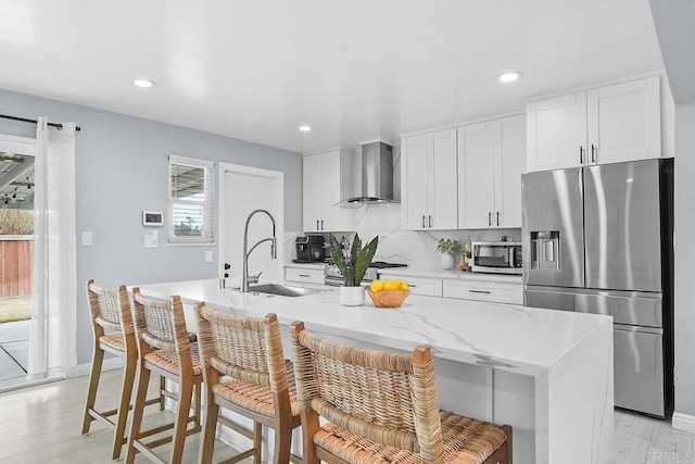 kitchen with sink, a breakfast bar area, stainless steel appliances, a center island with sink, and wall chimney range hood