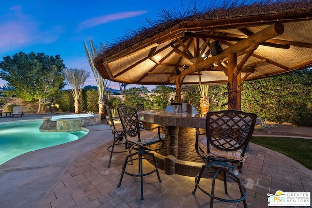 patio terrace at dusk with a pool with hot tub, a gazebo, exterior bar, and area for grilling