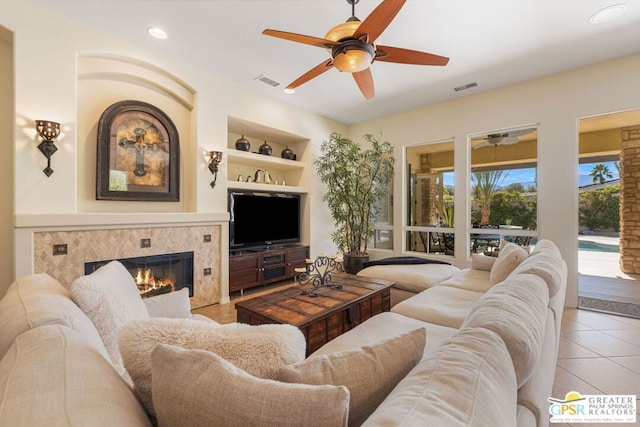 living room with a tile fireplace, built in features, light tile patterned floors, and ceiling fan