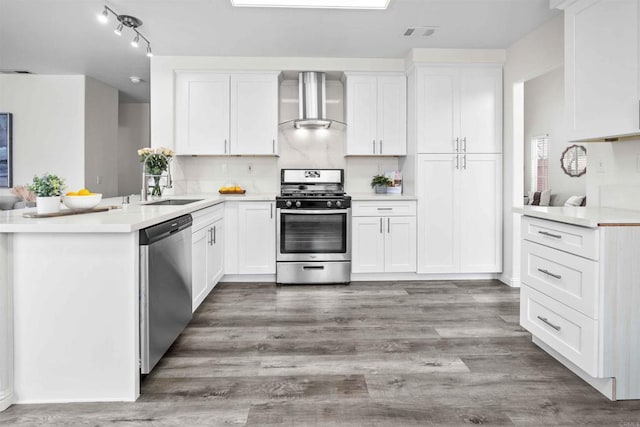 kitchen featuring appliances with stainless steel finishes, wood-type flooring, white cabinetry, kitchen peninsula, and wall chimney exhaust hood