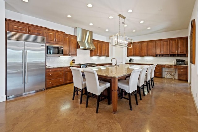 kitchen featuring decorative light fixtures, a kitchen breakfast bar, a kitchen island with sink, built in appliances, and wall chimney exhaust hood