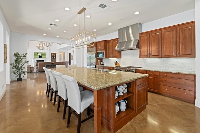 kitchen with sink, built in appliances, hanging light fixtures, a center island with sink, and wall chimney range hood