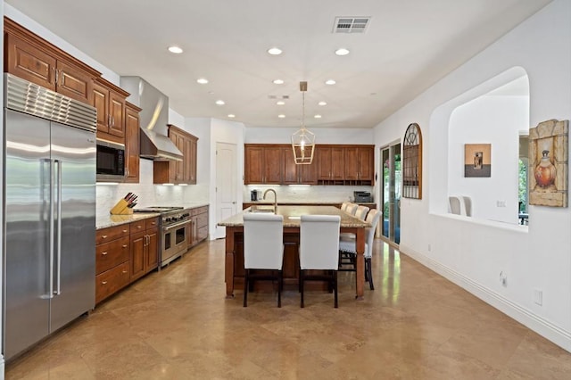 kitchen featuring wall chimney exhaust hood, a kitchen bar, built in appliances, light stone countertops, and a kitchen island with sink