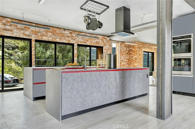 interior space with brick wall, island range hood, a kitchen island, light hardwood / wood-style flooring, and double oven