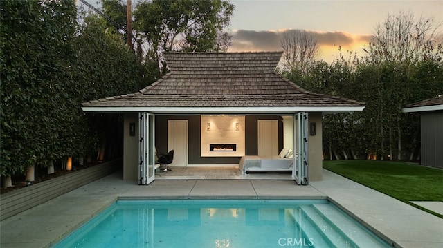 pool at dusk featuring an outdoor fireplace, a patio area, a lawn, and an outbuilding