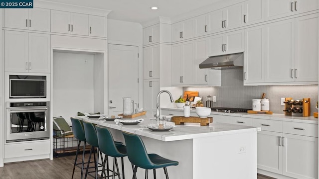 kitchen with white cabinetry, appliances with stainless steel finishes, a center island with sink, and exhaust hood