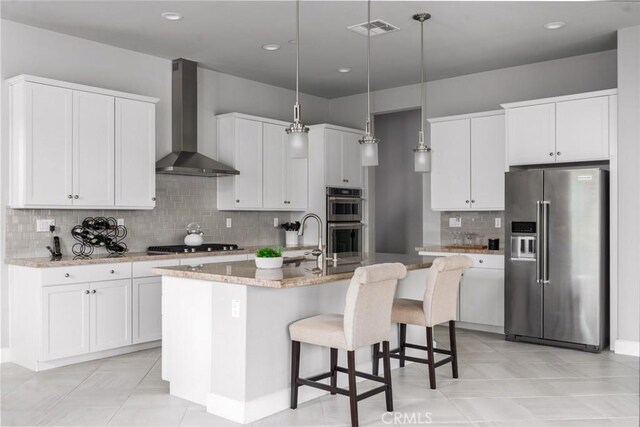 kitchen with decorative light fixtures, white cabinets, wall chimney range hood, a center island with sink, and black appliances