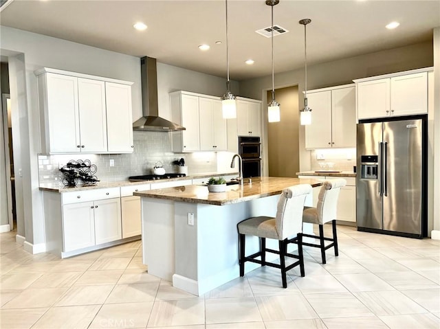 kitchen with wall chimney range hood, black appliances, white cabinets, a center island with sink, and decorative light fixtures