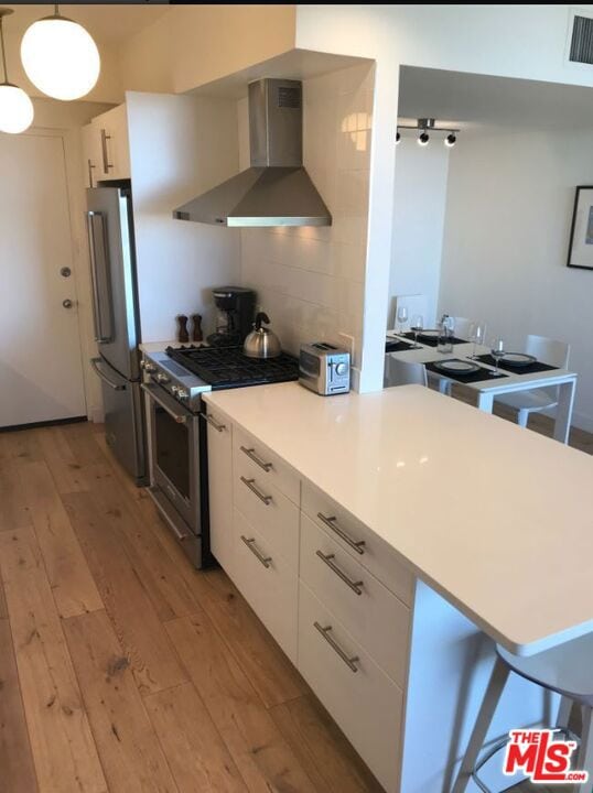 kitchen featuring premium appliances, wall chimney exhaust hood, white cabinetry, hanging light fixtures, and light hardwood / wood-style flooring