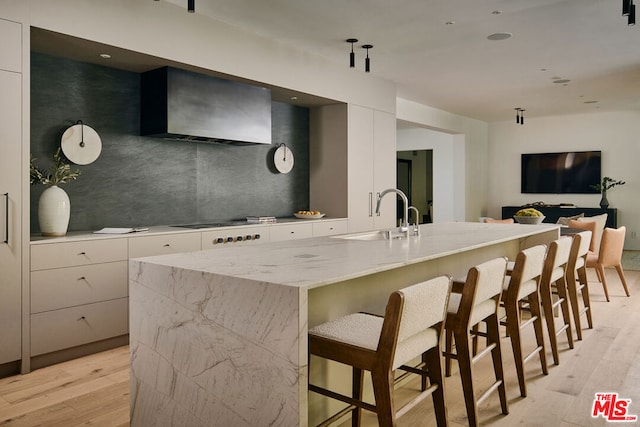 kitchen with sink, white cabinetry, wall chimney range hood, and a kitchen island with sink