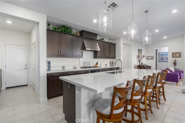 kitchen with hanging light fixtures, sink, dark brown cabinetry, wall chimney range hood, and a center island with sink