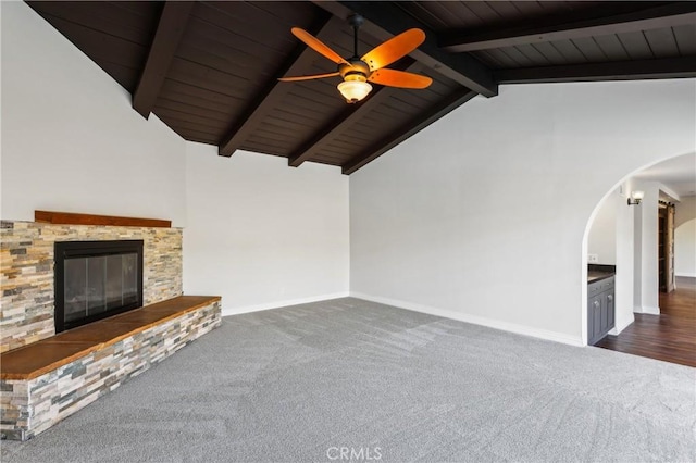 unfurnished living room with lofted ceiling with beams, ceiling fan, a stone fireplace, and dark carpet