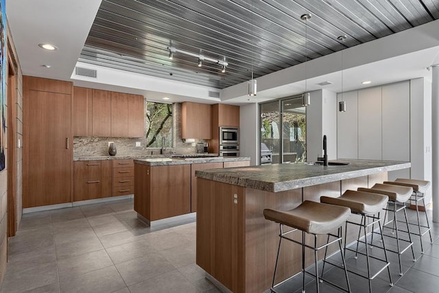 kitchen featuring decorative light fixtures, stainless steel appliances, sink, backsplash, and a center island with sink