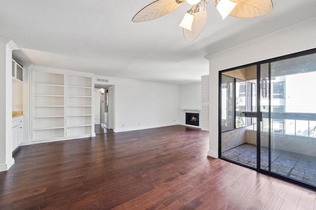 unfurnished living room with a ceiling fan, ornamental molding, dark wood-style flooring, a lit fireplace, and built in shelves
