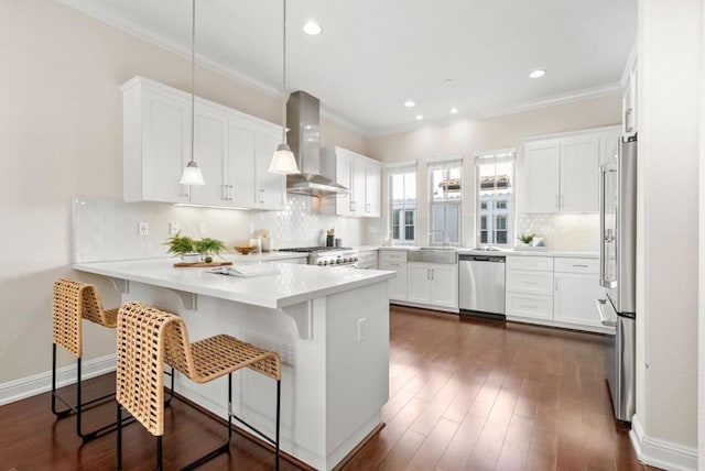 kitchen featuring wall chimney range hood, stainless steel appliances, a kitchen breakfast bar, white cabinets, and decorative light fixtures